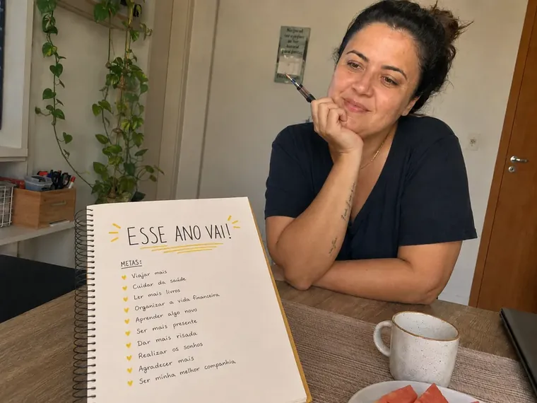 Mulher à mesa com um caderno de metas aberto, queixo apoiado na mão, olhar distante