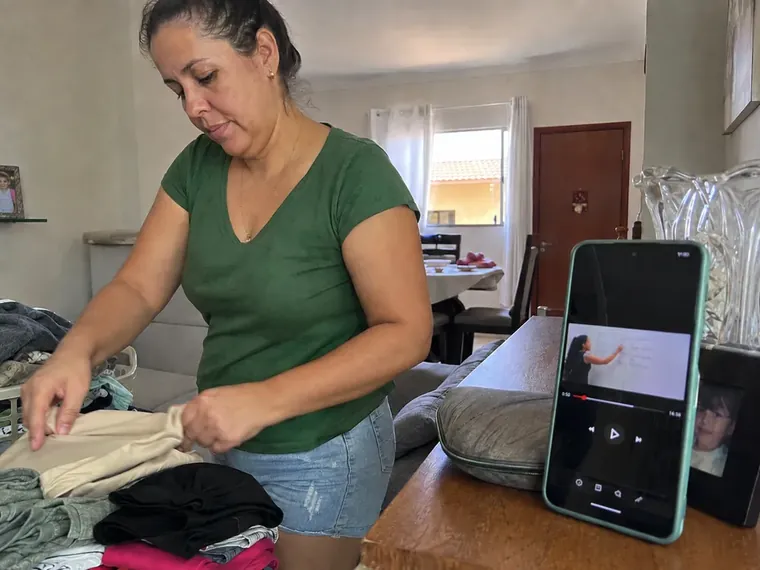 Mulher dobrando roupa enquanto uma aula toca no celular apoiado sobre a mesa