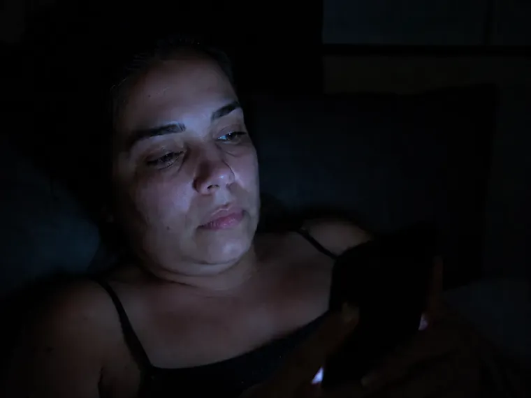Mulher deitada no escuro com o rosto iluminado pela tela do celular, expressão vazia